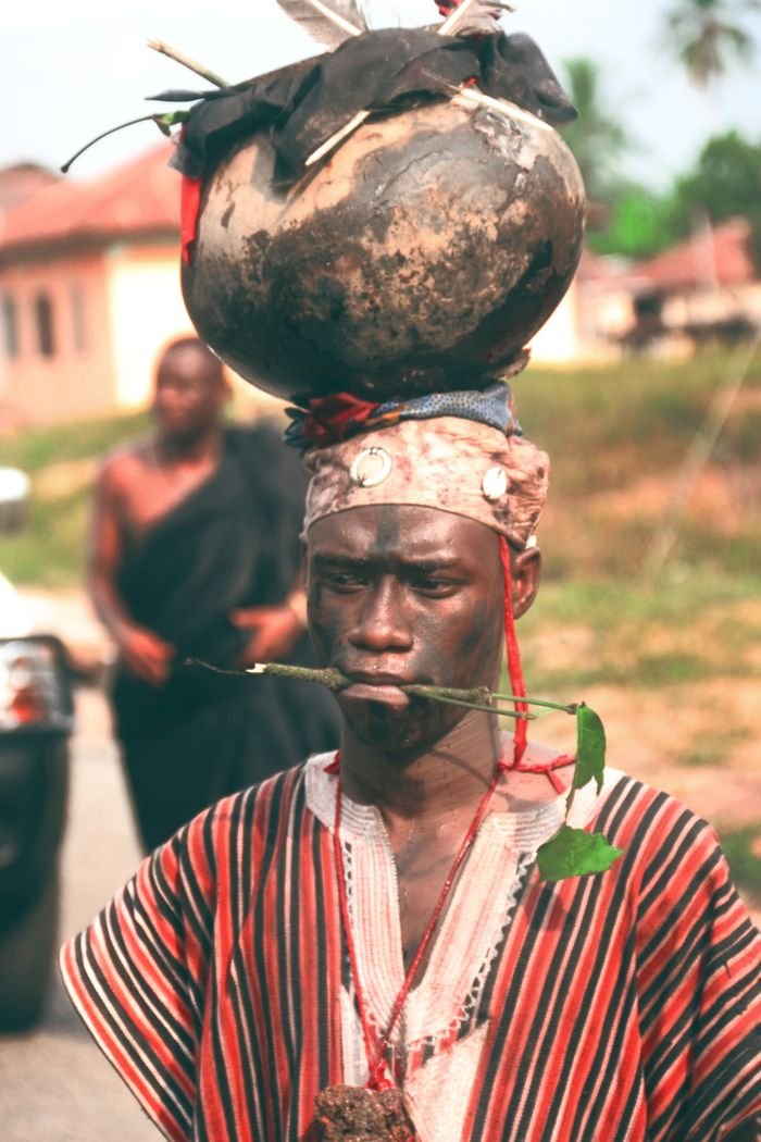About A Ghanaian man in traditional attire balances a pot on his head in a cultural display.